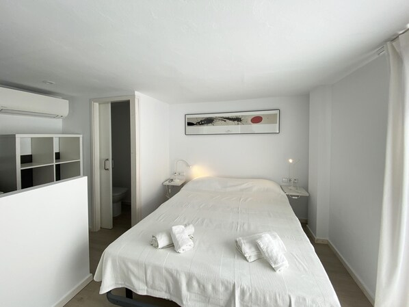 A hotel room with a bed, a bedside table with a lamp, a wall-mounted artwork, and a storage unit.