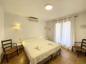 A hotel room with a bed, two chairs, a small table, an air conditioner, and a window with curtains.