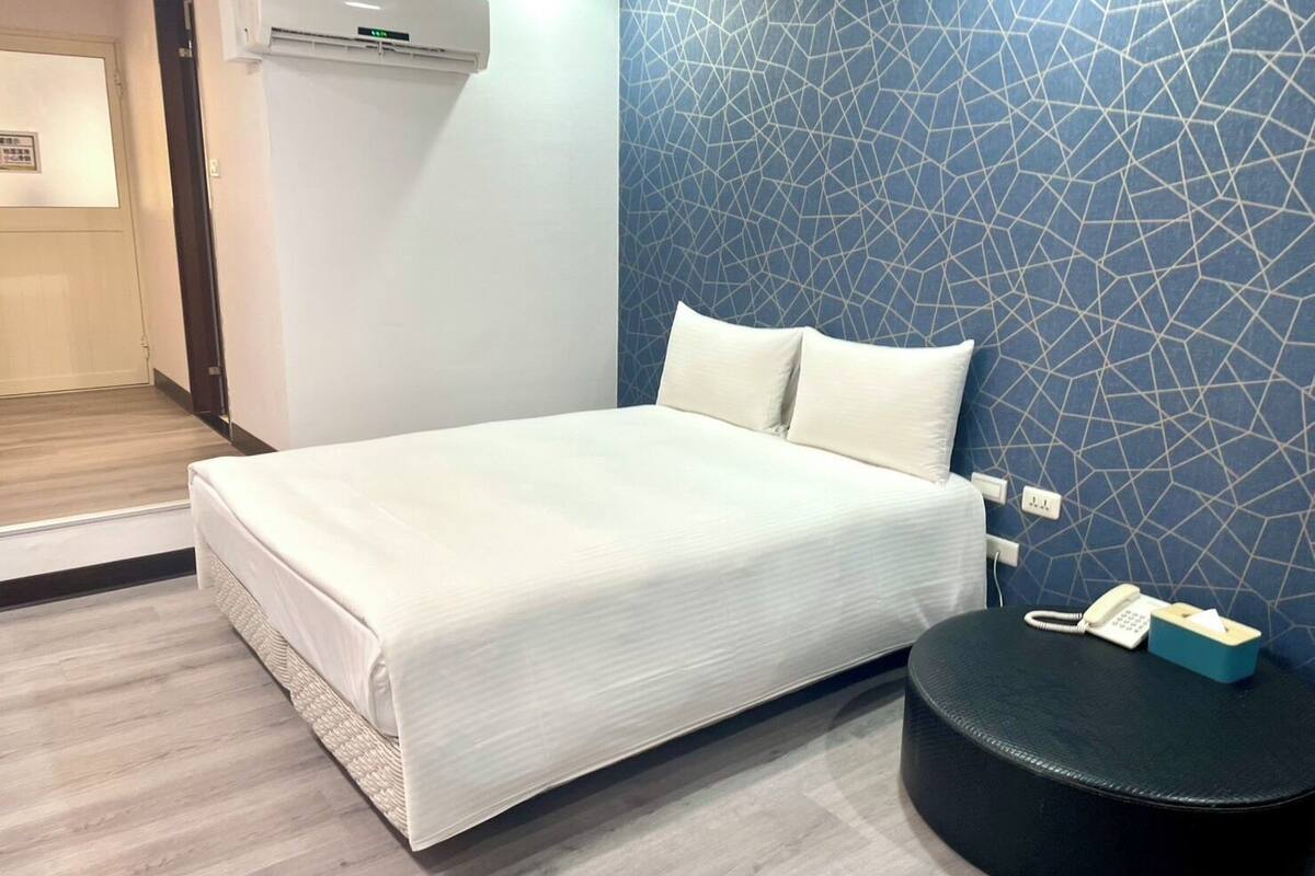 A hotel room with a bed, a wall-mounted phone, and a small black ottoman.