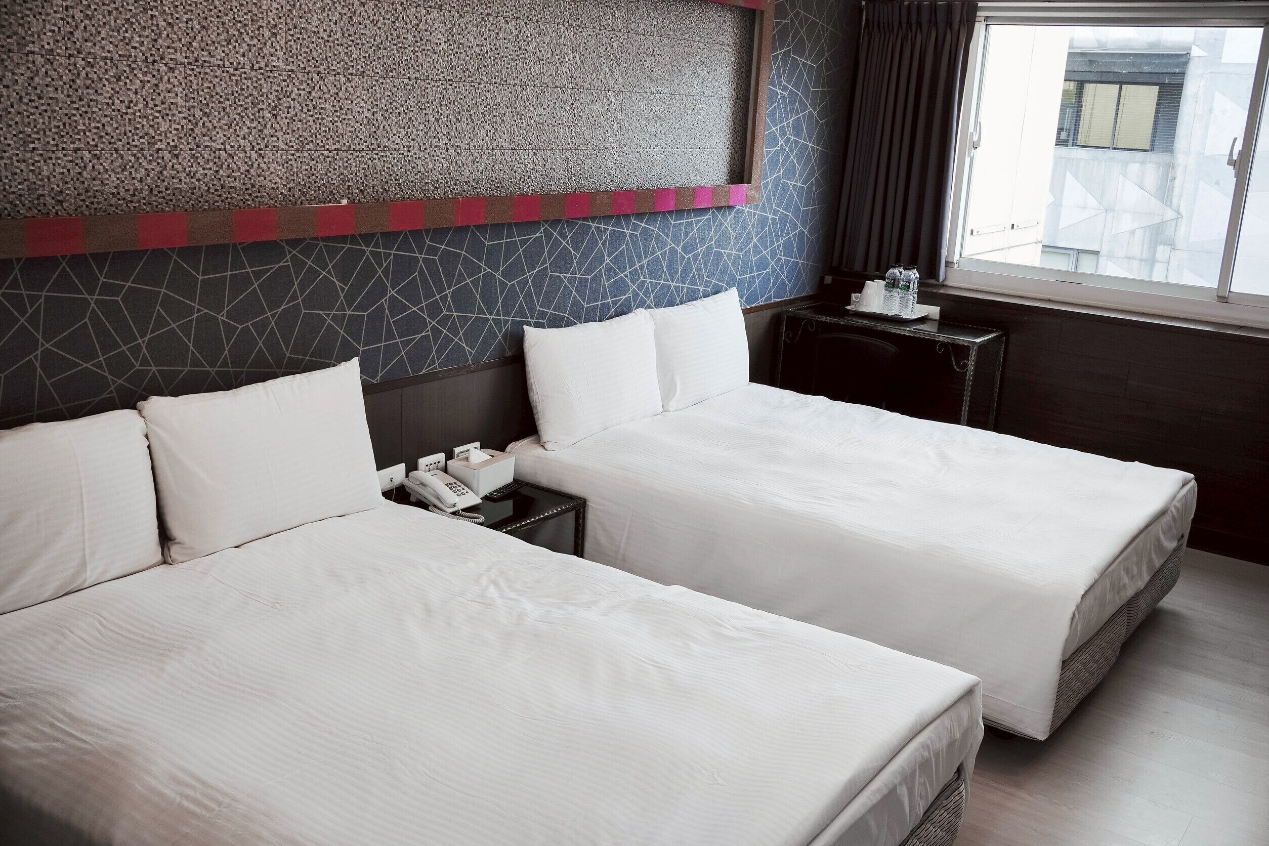 A hotel room with two beds, a small bedside table with a phone and tissues, and a window with a view of buildings.