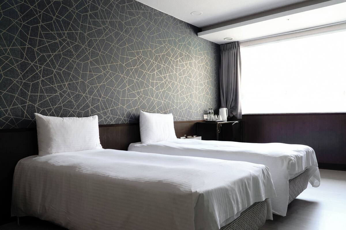 A hotel room with two beds, a geometric-patterned wall, a large window with curtains, and a small table with a lamp.