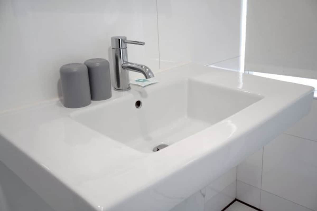 Un lavabo blanc dans une salle de bain, doté d’un robinet moderne et de deux récipients cylindriques, probablement des distributeurs de savon et de lotion, situés à gauche.