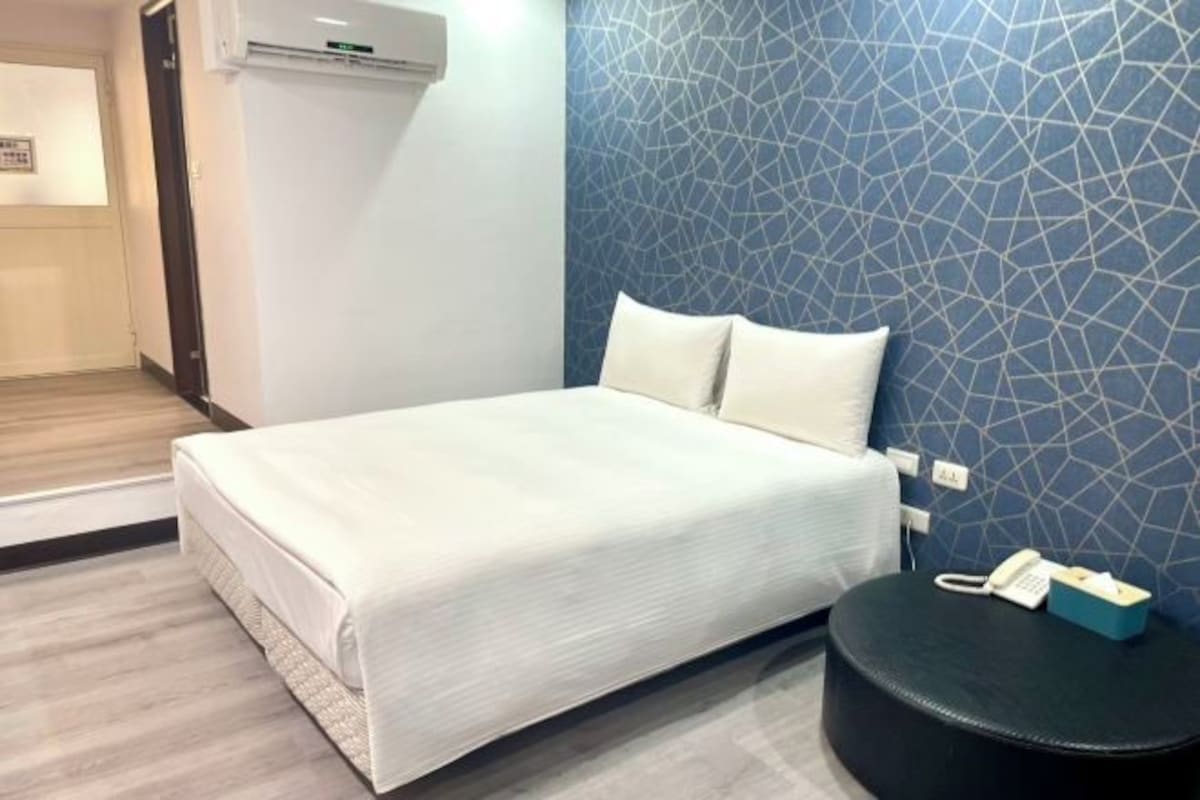 Une chambre d’hôtel avec un lit, un téléphone fixé au mur et une petite table sur laquelle se trouve une boîte de mouchoirs.