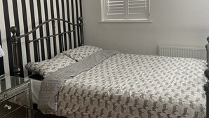 Een slaapkamer met een bed, een dressoir, een radiator, een raam met jaloezieën en een gestreept behang.