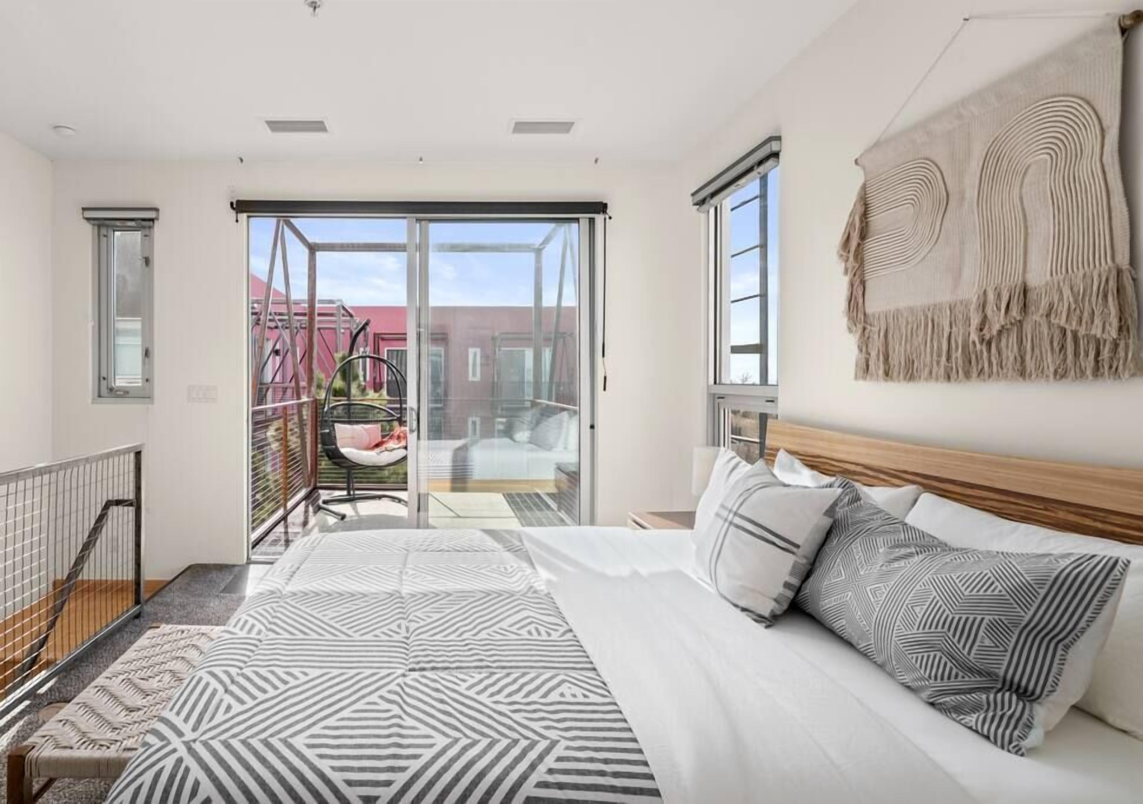 A modern bedroom with a large bed, a balcony with a chair, and a view of buildings outside.