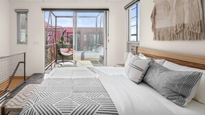 Une chambre moderne avec un grand lit, un balcon avec un fauteuil et une vue sur les bâtiments environnants.