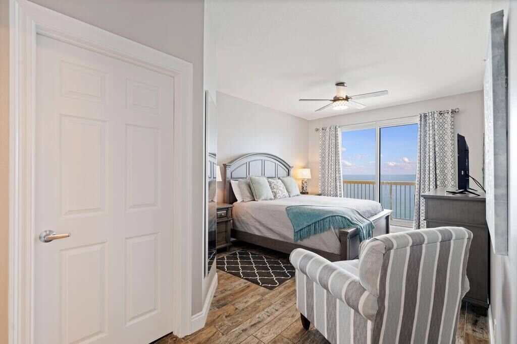 A bedroom with a bed, a chair, a desk, a ceiling fan, and a view of the ocean.