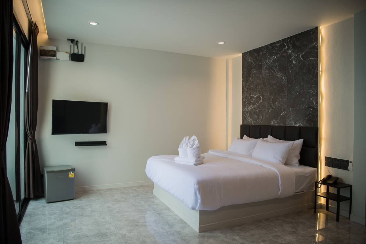 A modern hotel room with a large bed, a flat-screen TV mounted on the wall, a small bedside table with a lamp, and a marble-patterned wall.