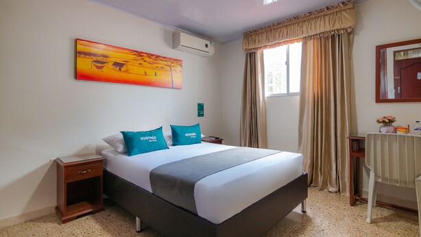 Une chambre d’hôtel avec un grand lit, une table de chevet, une commode, une fenêtre avec des rideaux, un tableau au mur et un appareil de climatisation.
