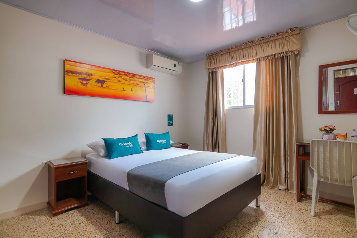 A hotel room with a large bed, a nightstand, a bedside table, a window with curtains, a painting on the wall, and an air conditioning unit.