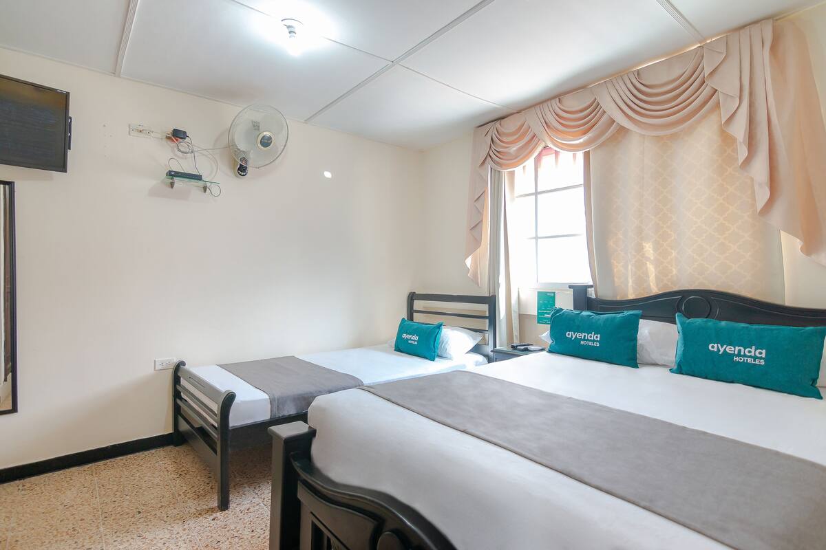 A hotel room with two beds, a TV mounted on the wall, a fan, and a window with curtains.