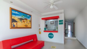 Un hall d'hôtel avec un canapé rouge, un tableau représentant une scène de plage, un ventilateur de plafond et un panneau indiquant « Ayenda Hotels ».