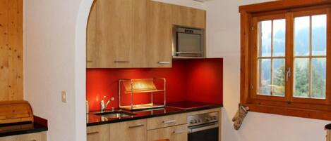 A compact kitchen with wooden cabinets, a microwave, a red backsplash, and a window with a view.