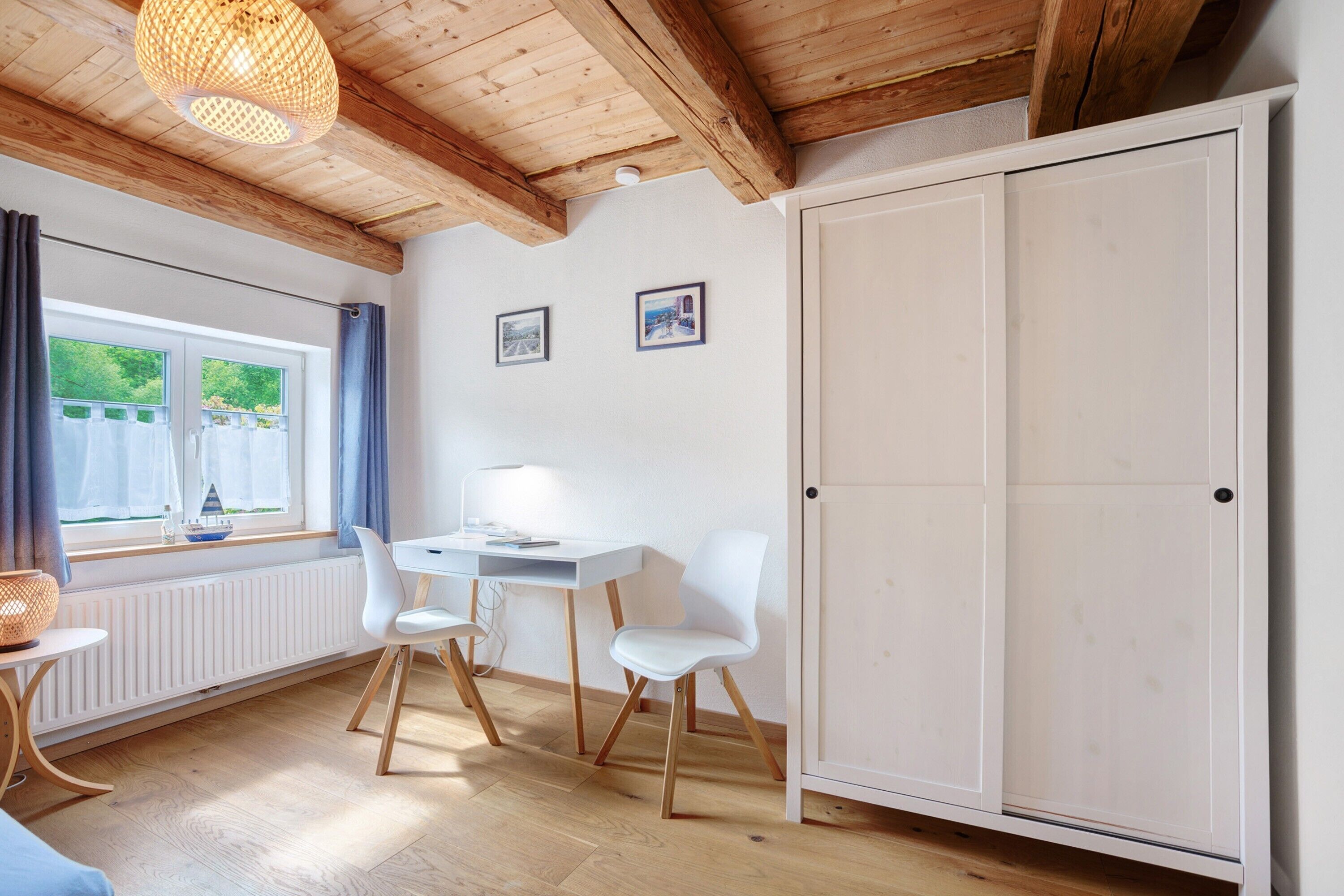 Una habitación con techo de madera, un radiador blanco, un escritorio blanco con dos sillas, un armario blanco y dos cuadros en la pared.