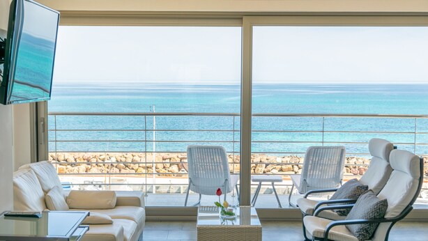 Ein modernes Wohnzimmer mit großem Fenster mit Blick auf eine felsige Küstenlinie und das Meer. Zwei weiße Sessel, eine Couch und ein kleiner Tisch mit Blumenvase sind zu sehen.