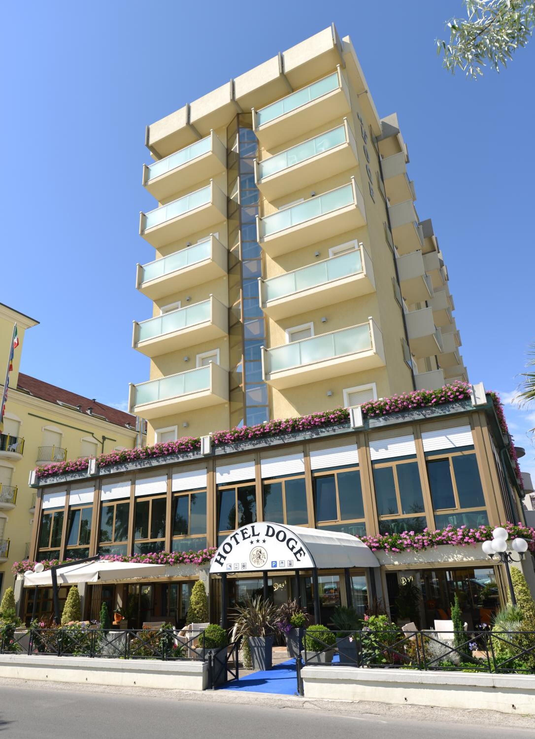 Un hotel a più piani con una segnaletica ben visibile che recita "HOTEL DOGE". Presenta balconi con ringhiere in vetro, una zona relax all'aperto coperta da ombrelloni e un cielo azzurro sullo sfondo.