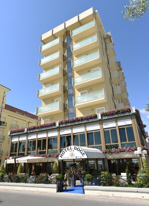 A multi-story hotel with a prominent sign reading "HOTEL DOGE". It features balconies with glass railings, a covered outdoor seating area with umbrellas, and a clear blue sky in the background.