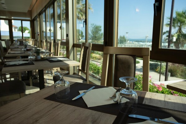 Comedor con mesas dispuestas para dos, ventanas amplias con vistas a palmeras y un paisaje lejano.