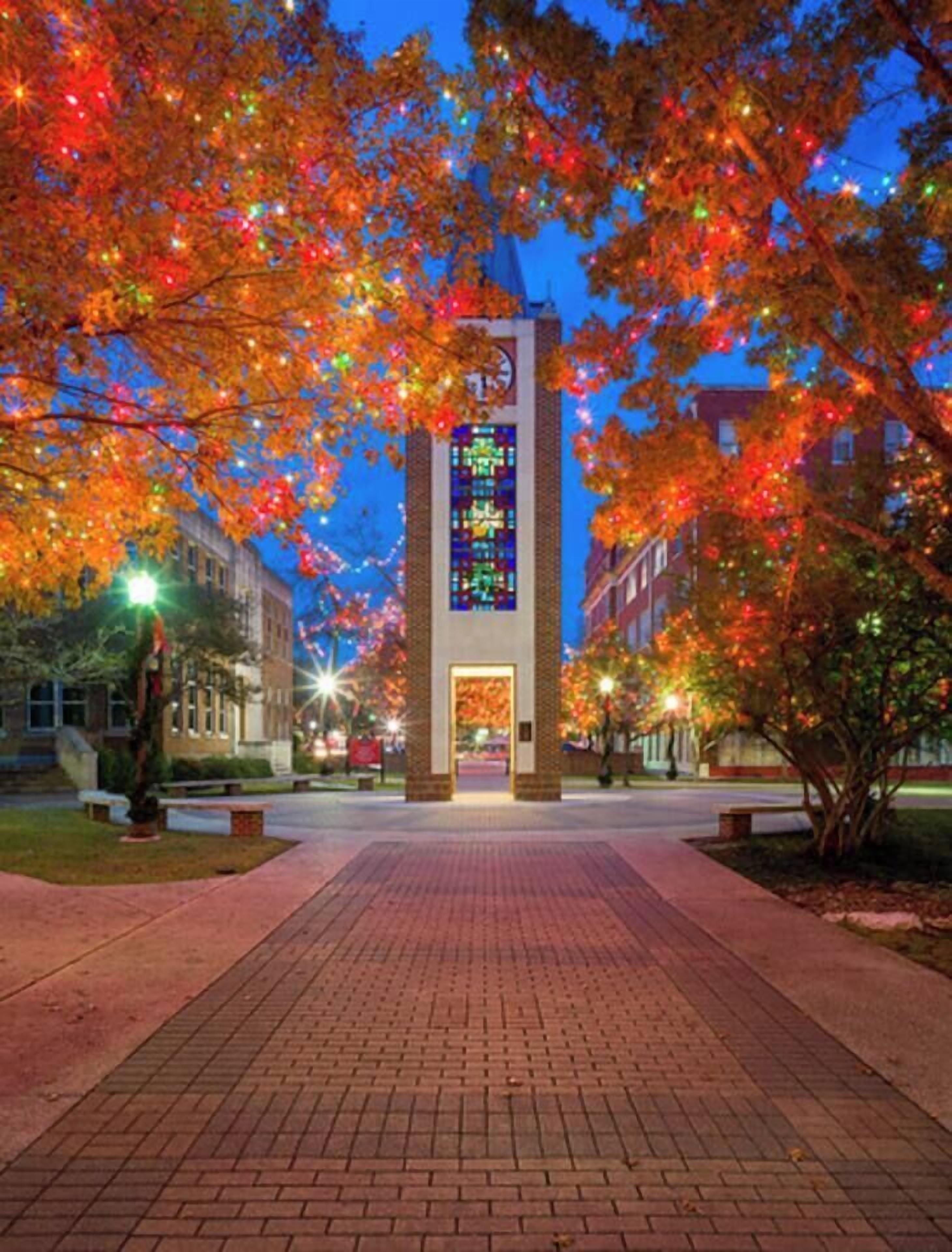 A brick pathway leading to a building with a stained glass window, surrounded by trees with colorful lights.