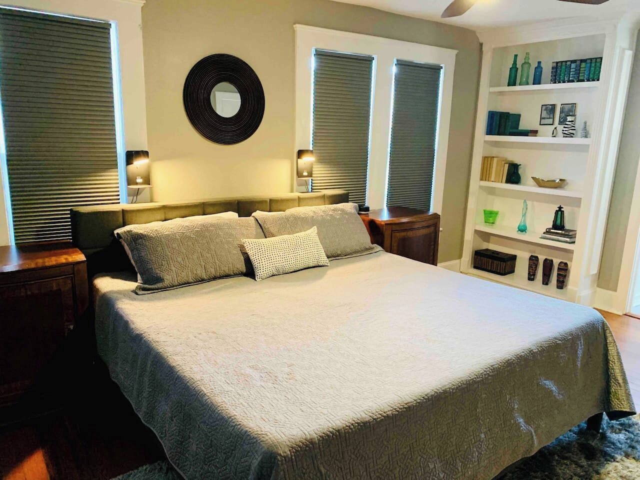 A bedroom with a bed, two bedside tables, a wall-mounted circular mirror, and a bookshelf.