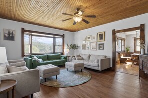 Ein geräumiges Wohnzimmer mit Holzbalkendecke, großen Fenstern und Blick ins Freie. Es gibt mehrere Sitzbereiche mit einer Kombination aus Sofas und Sesseln.
