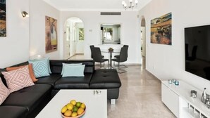A modern living room with a black sofa, a coffee table with fruit, and a dining area in the background.