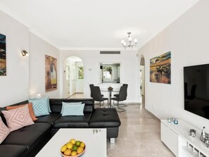 A modern living room with a black sofa, a coffee table with fruit, and a dining area in the background.