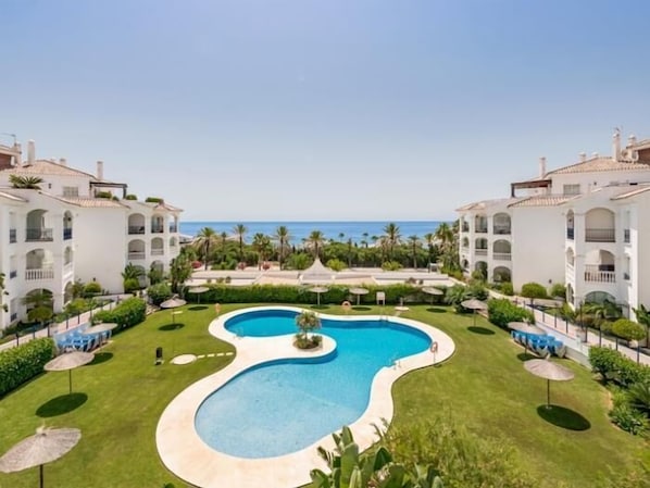 A resort with a central swimming pool, multiple buildings, and a view of the ocean.