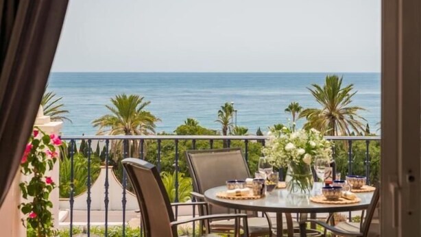 A balcony with a table set for dining, overlooking a calm sea and palm trees.