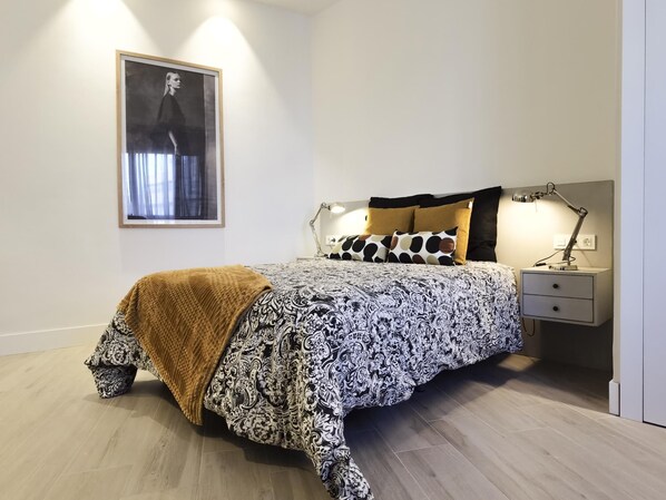 Une chambre à coucher avec un lit, une table de chevet, une lampe, un tableau encadré au mur et un plaid marron.