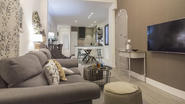 Ein modernes Wohnzimmer mit einem grauen Sofa, einem runden Hocker und einem an der Wand befestigten Flachbildfernseher.