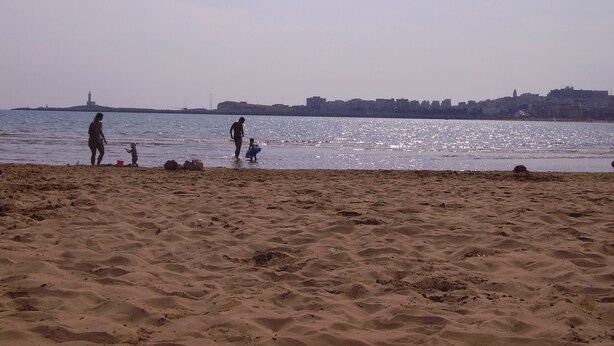 A beach with sand and water, a distant city skyline, and a lighthouse. - Holidays in Vieste in charming villas - 4 (Puglia)