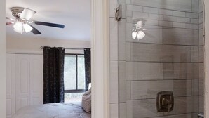 A bedroom with a bed, a ceiling fan, a window with curtains, and a bathroom with a shower and a towel rack.