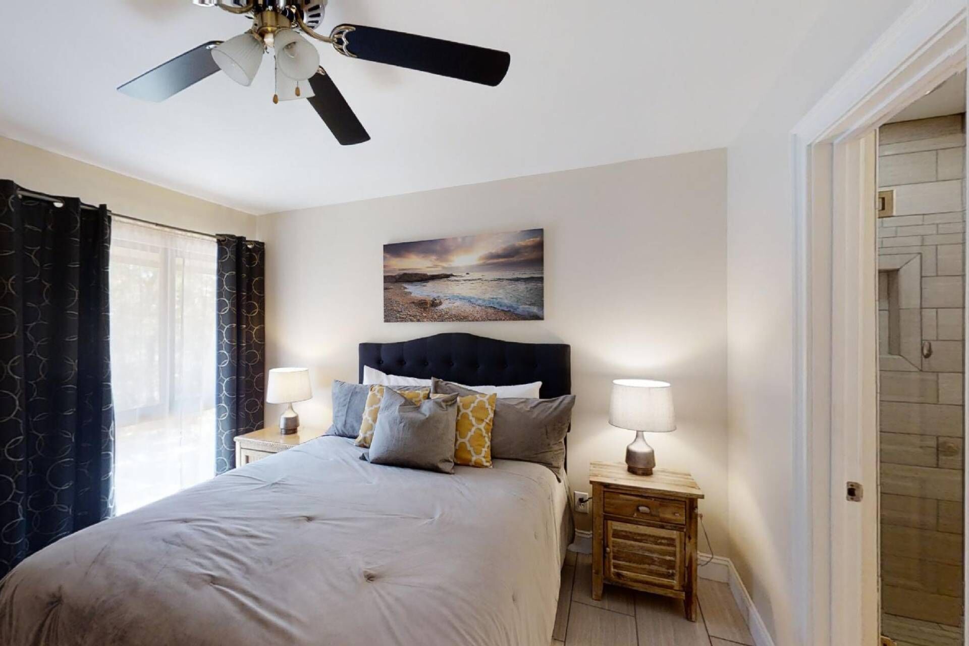 A bedroom with a bed, two bedside tables with lamps, a ceiling fan, a painting of a beach scene, and a door to the right.