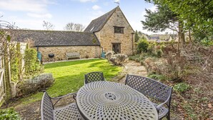 Une maison en pierre avec un toit en pente, une petite pelouse et une table en métal avec des chaises.