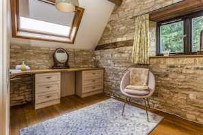 Cozy bedroom with sloped ceiling and exposed stone walls, featuring a desk, mirror, and plush armchair with a view of the surrounding greenery.