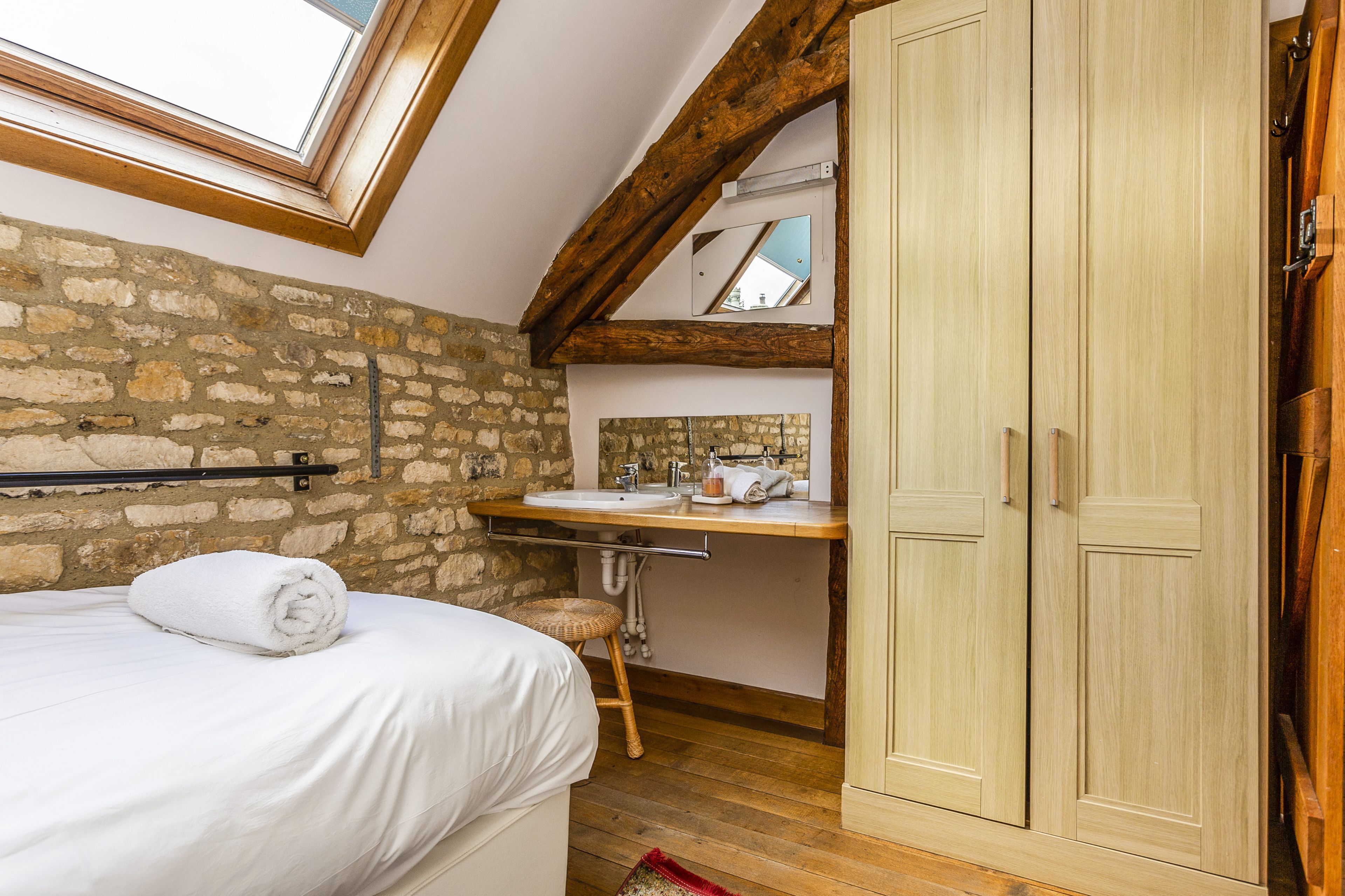 A bedroom with a stone wall, wooden beams, a skylight, a wooden bed, a white bathroom with a sink and mirror, and a wooden wardrobe.