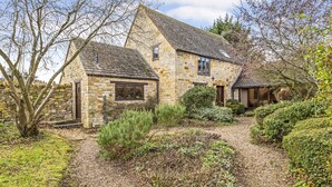 Une maison en pierre avec un toit en pente, un petit porche et un jardin où poussent différentes plantes, ainsi qu'un chemin de gravier.