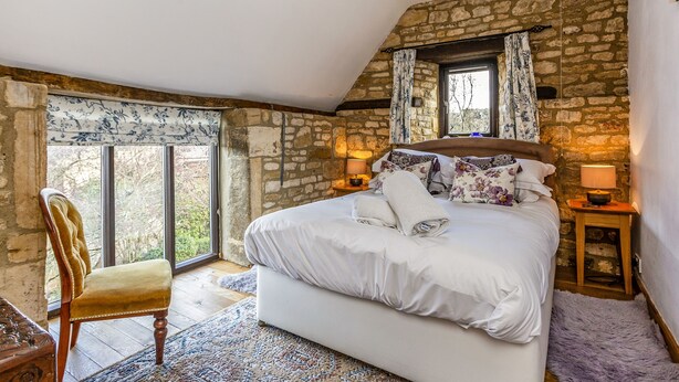 Une chambre avec un mur en pierre, un grand lit, une chaise en bois et une fenêtre avec des rideaux.