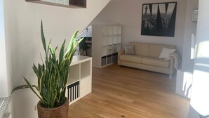A modern living room with a wooden floor, a beige sofa, a white shelving unit, and a large potted plant. - Modern 50 sqm apartment with living room, bedroom and balcony (Bad Aibling)