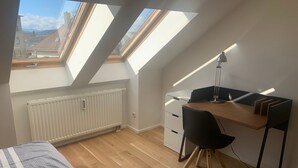 A small, modern room with a sloped ceiling, skylights, a wooden desk, a black chair, and a radiator. - Modern 50 sqm apartment with living room, bedroom and balcony (Bad Aibling)