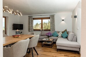 A modern living room with a wooden dining table, grey sofa, and a TV mounted on the wall.