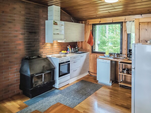 A compact kitchen with a brick wall, wooden cabinets, a built-in oven, a dishwasher, and a window with a view of greenery.