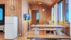 A wooden cabin interior with a dining table, benches, a refrigerator, a television, and a clock on the wall.
