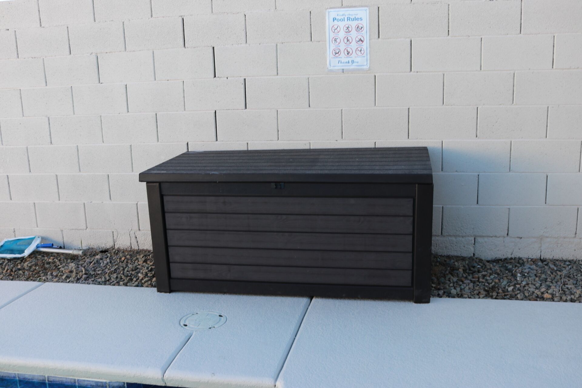 Une boîte de rangement de couleur sombre, placée contre un mur de briques blanches, avec un panneau indiquant les règles  à respecter au bord de la piscine, au-dessus.