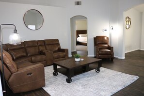 A living room with a brown leather sectional sofa, a brown leather armchair, and a coffee table with wheels. There is a round mirror on the wall and a floor lamp. - Beautiful oasis !!!! 10 minutes to the EDC festival  (North Las Vegas)