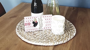 Un tavolo di legno con un sottopiatti intrecciato, una bottiglia di vino, una candela bianca, una scatola decorativa e un vaso con una pianta.