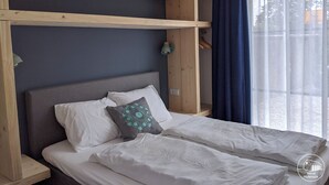 Un letto con biancheria bianca, una testata grigia e un cuscino decorativo. Sopra il letto si trova una mensola in legno e una lampada a sospensione.