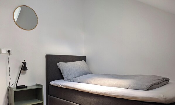 A single bed with a grey headboard and pillows, a bedside table with a lamp, and a wall-mounted mirror. - Nieuw Molenwijk, cozy holiday home  4-5 pers.
( Friday to Friday ) (Oostkapelle)