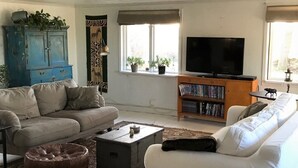 Ein Wohnzimmer mit einem beigefarbenen Sofa, einem weißen Sofa, einem Couchtisch, einer blauen Kommode mit Pflanzen, einem Bücherregal und einem Fernseher.